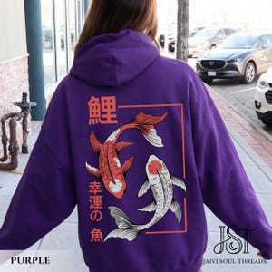 May include: A purple hoodie featuring a koi fish design. The back of the hoodie displays two koi fish in red and white, with Japanese text above and below the fish. The hoodie has a drawstring hood and long sleeves.