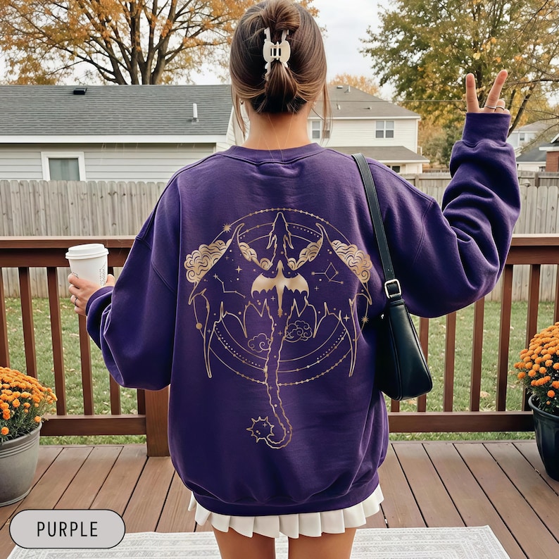 May include: A purple sweatshirt with a gold graphic design on the back. The design features a stylized dragon with wings, clouds, and constellations. The word "PURPLE" is in a white rectangle at the bottom.