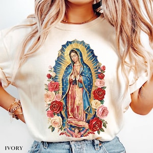 May include: Ivory-colored t-shirt featuring a religious illustration of a woman in a blue cloak, surrounded by roses. The illustration has a gold halo and is set against a blue background. The word "IVORY" is printed at the bottom.