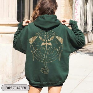 May include: Forest green hoodie with a gold dragon design on the back. The dragon is surrounded by a circular pattern with stars and clouds. The hoodie has a hood and long sleeves. The text "FOREST GREEN" is at the bottom.