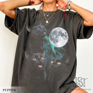 May include: A charcoal grey short-sleeve t-shirt featuring a graphic print of three black cats and a full moon against a green nebula background. The shirt has a crew neckline and a relaxed fit. The text JST Jaivi Soul Threads is visible.