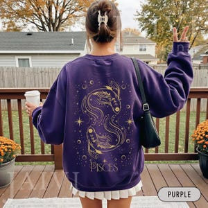 K&ouml;nnte beinhalten: Ein lila Sweatshirt mit einem goldenen Fische-Sternzeichen-Design auf dem R&uuml;cken. Das Design umfasst zwei Fische, Sterne und das Wort "PISCES". Das Sweatshirt ist einfarbig lila und hat lange &Auml;rmel.