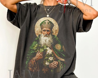 Puede incluir: Camiseta gris oscuro con una ilustración detallada de un santo con aureola, vistiendo una túnica verde y dorada, y sosteniendo flores. El diseño incluye el texto "JAIVI designs" en blanco.