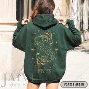 K&ouml;nnte beinhalten: Forstgr&uuml;ner Kapuzenpullover mit goldenem Fische-Design auf dem R&uuml;cken. Das Design umfasst zwei stilisierte Fische, Sterne und das Wort "PISCES". Der Hoodie hat eine Kapuze mit Kordelzug und eine K&auml;ngurutasche.
