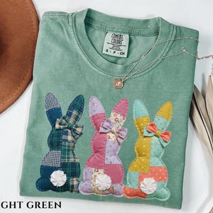 May include: Light green t-shirt with three patchwork bunny rabbit appliques. Each bunny has a different pattern and color, including plaid, floral, and solid colors. The shirt has a crew neck and short sleeves. The text "LIGHT GREEN" is at the bottom.