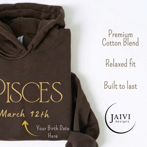 K&ouml;nnte beinhalten: Ein brauner Kapuzenpullover mit dem Wort "PISCES" in goldfarbenen Buchstaben. Darunter steht "March 12th", mit einem Pfeil, der auf "Your Birth Date Here" zeigt. Der Hoodie zeigt auch den Text "Premium Cotton Blend", "Relaxed fit" und "Built to last".