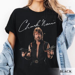 May include: Black t-shirt featuring a portrait of Chuck Norris holding two guns, with his signature in white script above the image. The word "BLACK" is printed in white at the bottom right corner of the shirt.