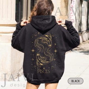 K&ouml;nnte beinhalten: Schwarzer Kapuzenpullover mit goldenem Fische-Sternzeichen-Design auf dem R&uuml;cken. Das Design zeigt zwei stilisierte Fische, Sterne und das Wort "PISCES". Der Hoodie hat eine Kapuze mit Kordelzug und lange &Auml;rmel.