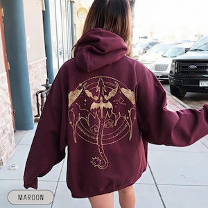 May include: Maroon hoodie with a gold dragon and constellation design on the back. The dragon is centered within a circular frame, with wings outstretched and a long tail. The word "MAROON" is printed at the bottom.