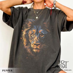 May include: A charcoal gray short-sleeve t-shirt featuring a large, detailed graphic print of a lion's face in shades of orange, blue, and black. The design includes subtle paint splatter effects. The text JST JAIVI SOUL THREADS is visible.