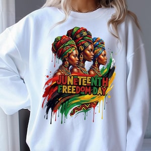 May include: White sweatshirt featuring a vibrant graphic of three women with colorful head wraps and jewelry. The design includes the text "Juneteenth Freedom Day" with red, black, green, and yellow accents. The artwork has a watercolor effect.
