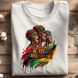 May include: White crewneck sweatshirt featuring a colorful graphic celebrating Juneteenth. The design includes three women wearing vibrant head wraps, with the words "Juneteenth Freedom Day" in bold letters. The artwork incorporates red, black, green, and yellow.