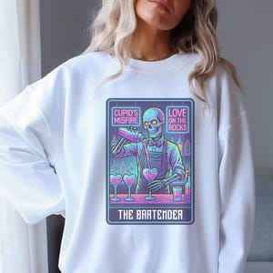 May include: White sweatshirt featuring a neon graphic of a skeleton bartender pouring drinks into heart-shaped glasses. The graphic includes the text "Cupid's Misfire," "Love on the Rocks," and "The Bartender."