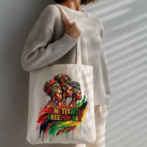 May include: A cream-colored tote bag featuring a vibrant illustration of three women wearing colorful head wraps. The design includes the text "Juneteenth Freedom Day" with a colorful paint stroke design.
