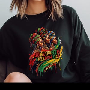 May include: Black sweatshirt featuring a colorful graphic of three women wearing head wraps, with the text "Juneteenth Freedom Day" in red, yellow, and green. The design incorporates the colors of the Pan-African flag.