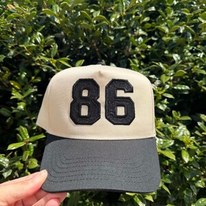 May include: A beige and black baseball cap with the number "86" in black, stitched on the front. The cap has a black brim and a structured, classic design. The background is a lush green bush.