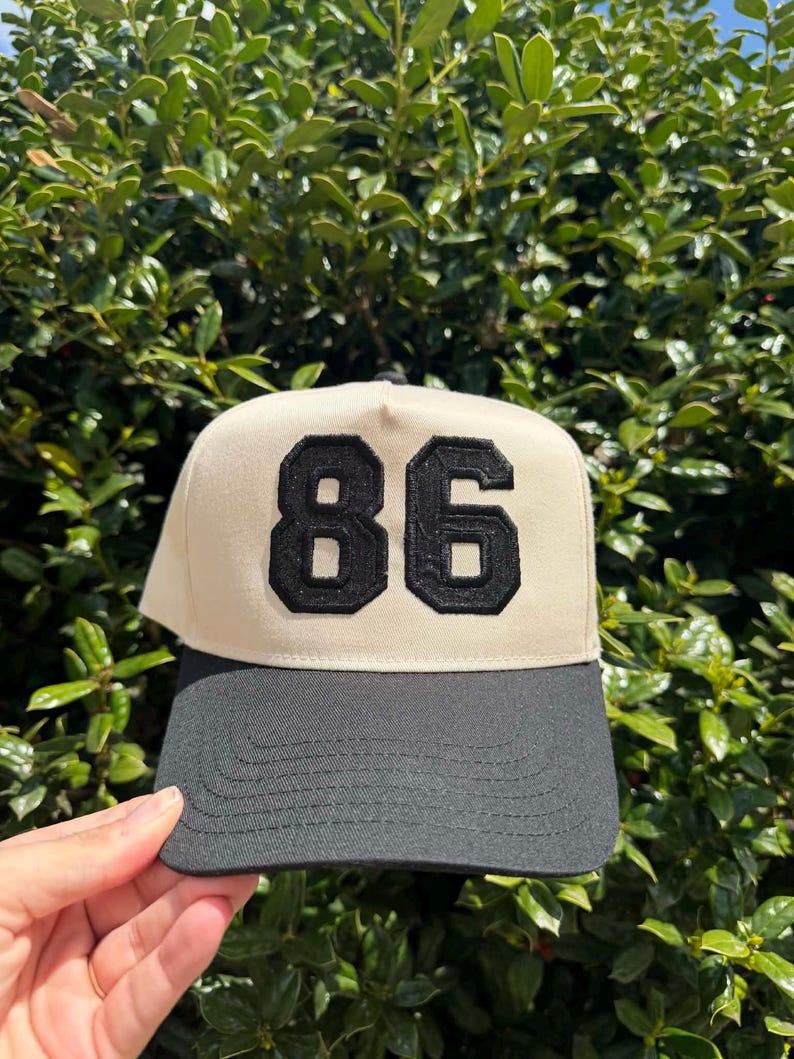 May include: A beige and black baseball cap with the number "86" in black, stitched on the front. The cap has a black brim and a structured, classic design. The background is a lush green bush.