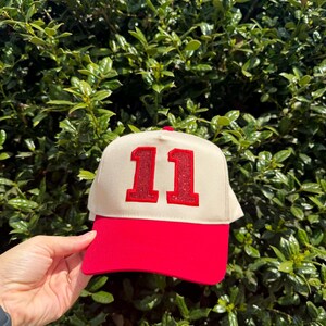 May include: A red and cream-colored baseball cap with the number "11" in red glitter. The cap has a red brim and a cream-colored front panel. The cap is set against a background of green foliage.