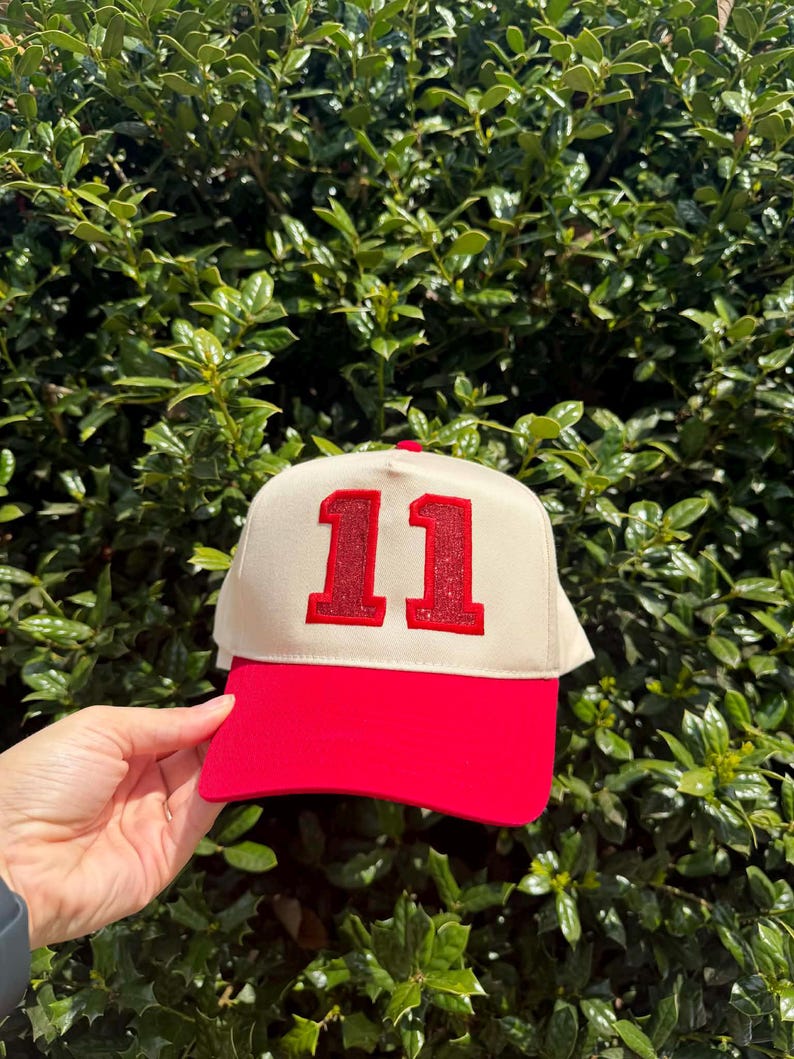 May include: A red and cream-colored baseball cap with the number "11" in red glitter. The cap has a red brim and a cream-colored front panel. The cap is set against a background of green foliage.