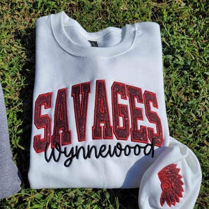 May include: White sweatshirt with red glitter lettering that says "SAVAGES Wynnewood" and a red glitter Native American headdress on the sleeve.