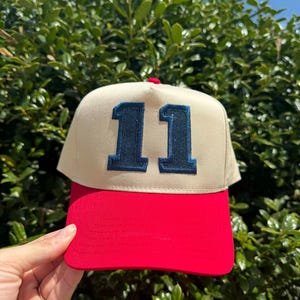May include: A beige and red baseball cap with the number "11" embroidered in navy blue on the front. The cap has a red brim and a red button on top. The background is a green bush.