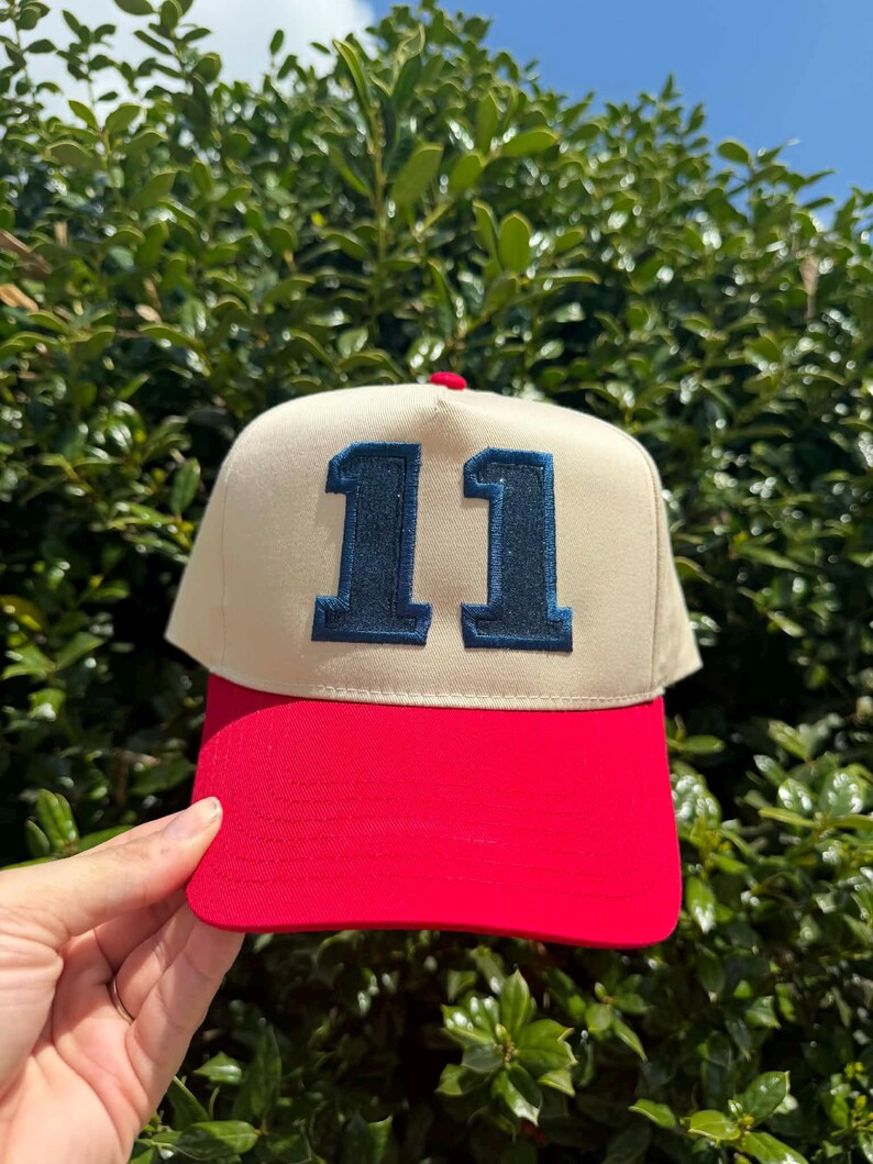 May include: A beige and red baseball cap with the number "11" embroidered in navy blue on the front. The cap has a red brim and a red button on top. The background is a green bush.