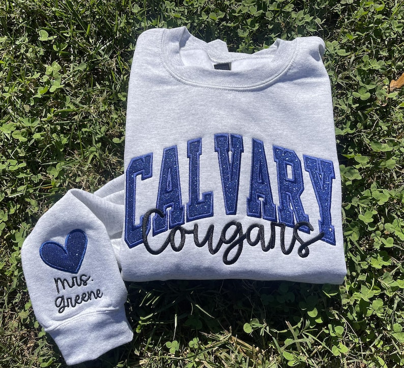 May include: A gray sweatshirt with the text "CALVARY Cougars" in blue glitter lettering. A smaller gray sweatshirt with a blue heart and the text "Mrs. Greene" is also pictured.