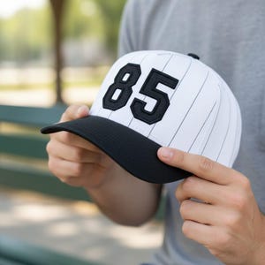 Pu&ograve; includere: Un cappellino da baseball bianco con sottili strisce nere e visiera nera. Il numero "85" &egrave; visualizzato in nero sul davanti. Il cappellino &egrave; tenuto da una persona.