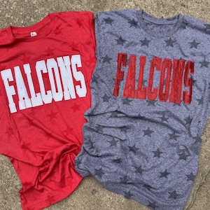May include: Two t-shirts, one red and one grey, each with a star pattern. The word "FALCONS" is printed on each shirt in large block letters. The red shirt has white lettering, while the grey shirt has red lettering.