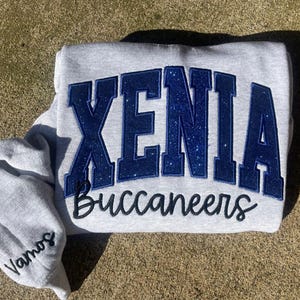 May include: Gray sweatshirt with blue glitter lettering that says "XENIA Buccaneers" in a large, arched font.