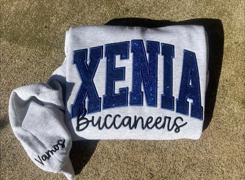 May include: Gray sweatshirt with blue glitter lettering that says "XENIA Buccaneers" in a large, arched font.