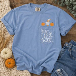 Puede incluir: Una camiseta de manga corta azul claro con un bolsillo en el pecho que presenta una oca blanca bordada con un lazo naranja, dos calabazas naranjas y el texto cursivo blanco It's fall y'all! La camiseta tiene cuello redondo y una etiqueta Comfort Colors.