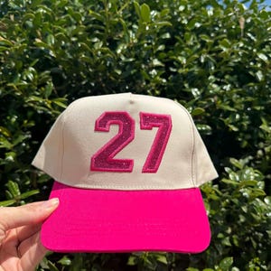 May include: A baseball cap with a cream-colored crown and a bright pink brim. The number "27" is prominently displayed on the front in a glittery pink font. The cap is held up against a backdrop of green foliage.