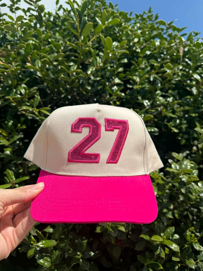 May include: A baseball cap with a cream-colored crown and a bright pink brim. The number "27" is prominently displayed on the front in a glittery pink font. The cap is held up against a backdrop of green foliage.