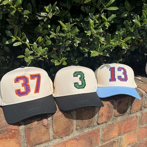 May include: Three baseball caps with cream-colored crowns and black or blue brims. Each cap features a different number: 37 in purple and orange, 3 in green, and 13 in red and blue. The caps are displayed on a brick wall.