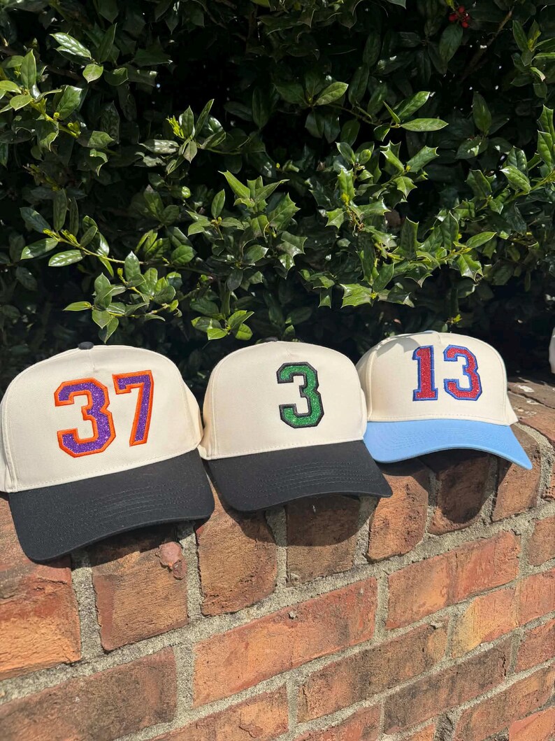 May include: Three baseball caps with cream-colored crowns and black or blue brims. Each cap features a different number: 37 in purple and orange, 3 in green, and 13 in red and blue. The caps are displayed on a brick wall.