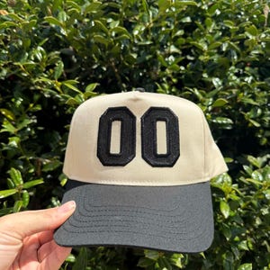 May include: A beige and black baseball cap with the number "00" in black on the front. The cap has a black brim and a black top button. The background is a green bush.