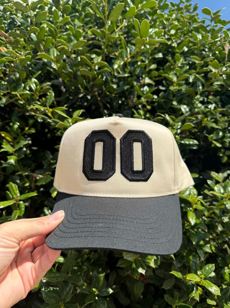 May include: A beige and black baseball cap with the number "00" in black on the front. The cap has a black brim and a black top button. The background is a green bush.