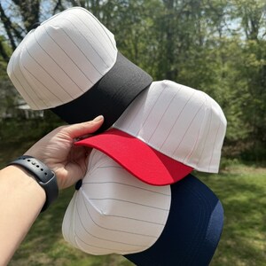 Pu&ograve; includere: Una pila di cappellini da baseball con tessuto bianco e sottili gessati neri. I cappellini hanno visiere di diversi colori: nero, rosso e blu navy. I cappellini sono tenuti contro uno sfondo di erba verde e alberi.
