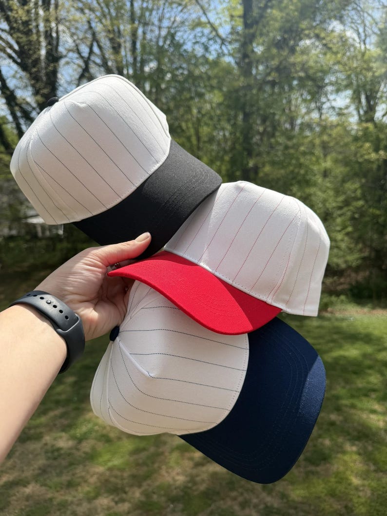 Pu&ograve; includere: Una pila di cappellini da baseball con tessuto bianco e sottili gessati neri. I cappellini hanno visiere di diversi colori: nero, rosso e blu navy. I cappellini sono tenuti contro uno sfondo di erba verde e alberi.
