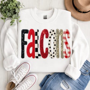 May include: A white long-sleeve sweatshirt featuring the word Falcons in large, decorative letters. Each letter has a unique pattern, including black sequins, red polka dots, black and white checkers, red sequins, black polka dots, gold glitter, and red plaid.
