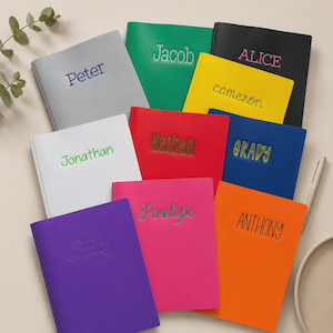 May include: A collection of eleven rectangular notebooks in various solid colors including gray, green, black, yellow, white, red, blue, purple, pink, and orange. Each notebook features a unique name embroidered or printed on the front cover.