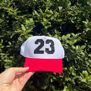 May include: A white baseball cap with a red brim and the number 23 in black. The cap is held up against a backdrop of green foliage, showcasing the hat's design and color contrast.