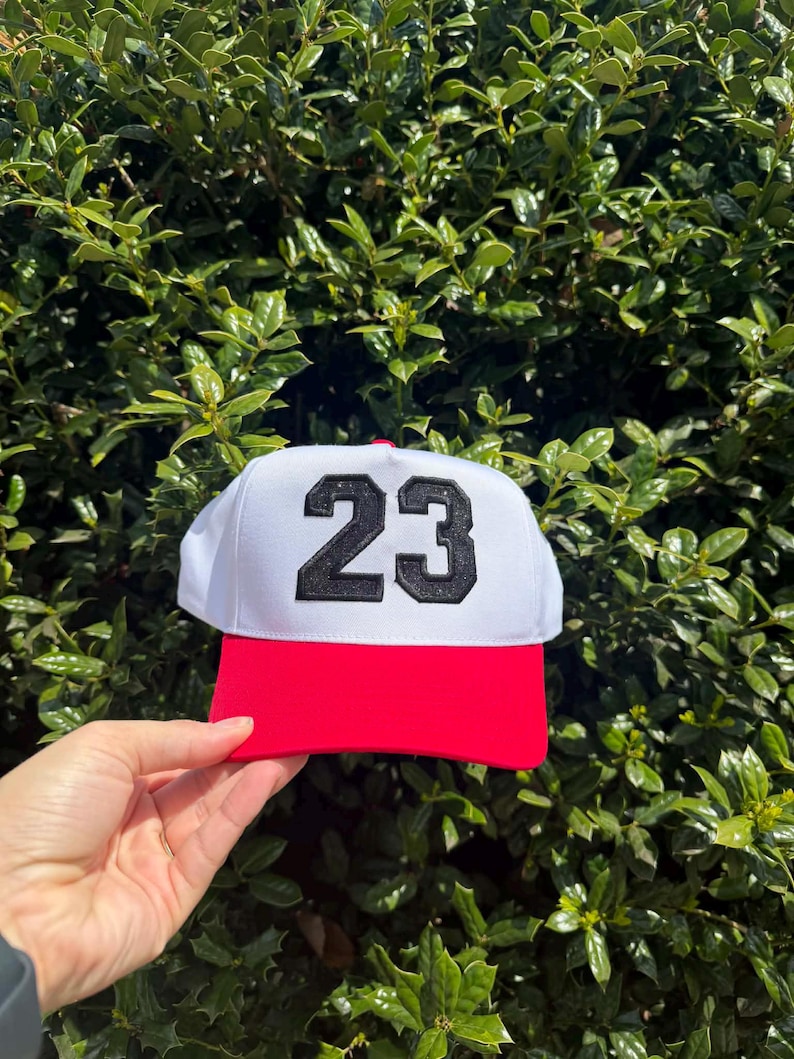 May include: A white baseball cap with a red brim and the number 23 in black. The cap is held up against a backdrop of green foliage, showcasing the hat's design and color contrast.