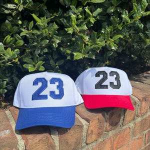 May include: Two baseball caps with the number 23 embroidered on the front. One cap has a white crown and blue brim with blue embroidery. The other cap has a white crown and red brim with black embroidery. The caps are displayed on a brick wall.