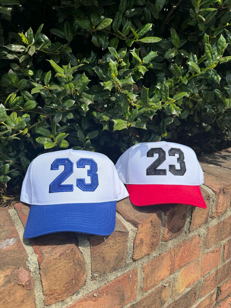 May include: Two baseball caps with the number 23 embroidered on the front. One cap has a white crown and blue brim with blue embroidery. The other cap has a white crown and red brim with black embroidery. The caps are displayed on a brick wall.