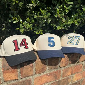 May include: Three baseball caps with cream-colored crowns and navy blue brims are displayed on a brick wall. Each cap features a different number: 14 in red glitter, 5 in blue glitter, and 27 in teal glitter. The caps are set against a backdrop of green foliage.