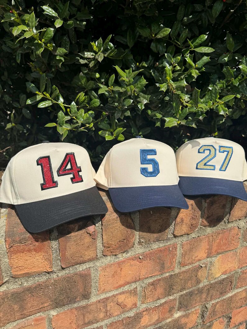 May include: Three baseball caps with cream-colored crowns and navy blue brims are displayed on a brick wall. Each cap features a different number: 14 in red glitter, 5 in blue glitter, and 27 in teal glitter. The caps are set against a backdrop of green foliage.
