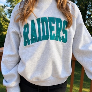 May include: A light gray crewneck sweatshirt featuring the word RAIDERS in large, dark teal, block-style embroidered letters across the chest. The garment has long sleeves, a ribbed mock neck collar, and ribbed cuffs and hem.
