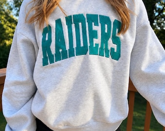 以下が含まれることがあります： 胸元にRAIDERSという文字がダークティールカラーの大きなブロック体で刺繍された、ライトグレーのクルーネックトレーナー。長袖で、リブ編みのモックネックカラー、袖口、裾が特徴的なデザインです。